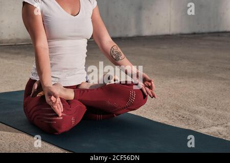 Eine nicht erkennbare Frau mit unbeteiltem Fuß, einem Tattoo in weißem Hemd und burgunderroten Hosen, die in einer Padmasana mit Gyan Mudra sitzt und auf der Straße zwischen der städtischen Umgebung gegen eine Betonwand meditiert Stockfoto