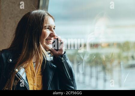 Junge lachende weibliche Reisende in lässiger Kleidung mit Koffer und Tasche steht am Fenster und lehnt sich an die Wand und spricht weiter Smartphone am Flughafen Stockfoto