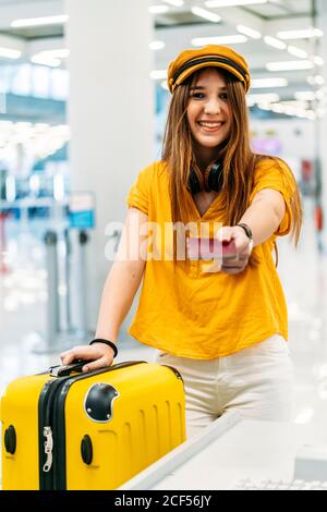 Fröhliche lächelnde Dame in bunten stilvollen Kleidung und Kopfhörer geben Pass und Blick auf die Kamera beim Check-in stehen Schalter im Flughafenterminal Stockfoto