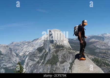 Seitenansicht des lässigen Mannes in Sonnenbrille mit Rucksack stehend Und Blick auf den felsigen Hügel an sonnigen Tag In den USA Stockfoto