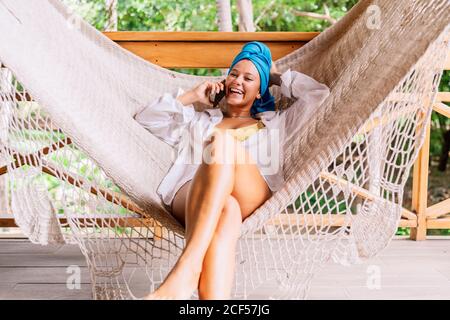 Fröhliche Frau in Hemd und hellem Kopftuch sitzt in der Hängematte und spricht auf dem Handy und schaut auf die Kamera von grünen Bäumen in Costa Rica Stockfoto