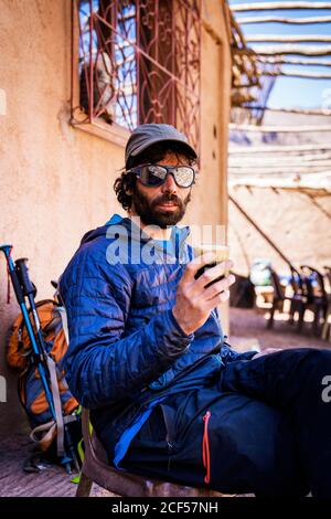 Entspannter Mann in warmer Kleidung und Sonnenbrille, während er Getränke trinkt Auf einem Stuhl neben dem Rucksack auf der sonnigen Straßenterrasse sitzen Stockfoto