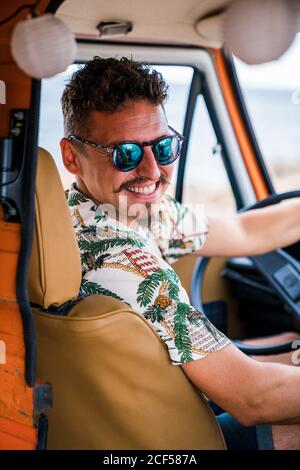 Stilvoller Schnurrbart Mann in Sonnenbrille und buntes Hemd halten Lenkung Rad und lächelnd auf Kamera im Auto am Strand Stockfoto