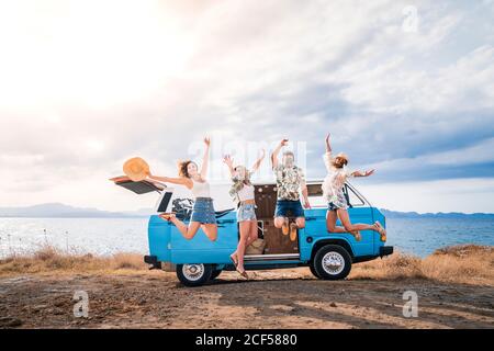 Fröhliche Tourist springen heben Hände und Beine auf dem Hintergrund Heller Minivan am Strand an sonnigen Tagen Stockfoto