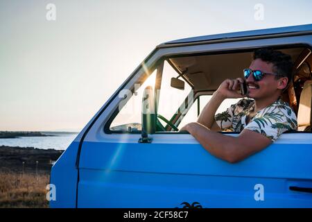 Seitenansicht des lächelnden erwachsenen Mannes in Sonnenbrillen und lässig Kleidung, die wegschaut und telefonieren kann, während man im Sitzen sitzt Blauer Van an sonniger Abendküste Stockfoto