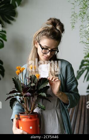 Junge Frau Floristin in Brillen tragen Leinenkleid, hält blühende calathea Pflanze in alten roten Milch kann in ihrem Garten zu Hause stehen.Liebe zur Pflanze Stockfoto