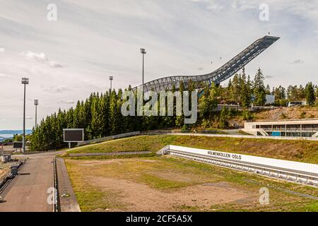 Der Holmenkollen-Skisprung erhebt sich über einer Zugangsstraße in der Nähe von Oslo, Norwegen, mit einem Schild „Holmenkollen Oslo“ im Vordergrund. Der aktuelle, etwa 60 Meter hohe Turm, der 2010 von den Architekten JDS/Julien De Smedt neu gebaut wurde, hat die Olympischen Winterspiele 1952 und mehrere Nordische Skiweltmeisterschaften ausgerichtet. Stockfoto