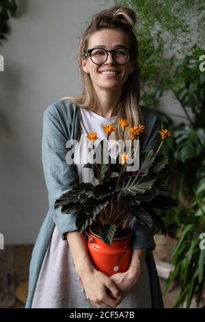 Glückliche Frau Gärtnerin in Brillen tragen Leinenkleid, hält eine blühende calathea Pflanze in alten roten Milch kann Blick auf Kamera, stehend in ihrem Haus Stockfoto