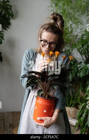 Junge lächelnde Frau Gärtnerin in Brillen tragen Leinenkleid, hält eine blühende calathea Pflanze in alten roten Milch kann in ihrem Garten zu Hause stehen Stockfoto