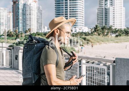 Profil, attraktiver junger Mann mit Rucksack, hält ein Smartphone, verschwommen Gebäude Hintergrund. Stockfoto