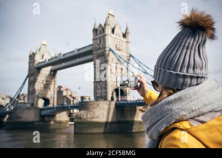 Rückansicht der weiblichen Reisenden in warmer Kleidung und Hut Fotografieren der Tower Bridge mit dem Smartphone während der Besichtigung in London an sonnigen Tagen Stockfoto
