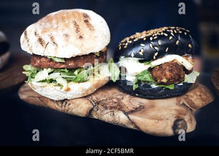 Leckere weiße und schwarze Burger mit grünem Salat und Käse Serviert auf Holzbrett auf schwarzem Tisch Stockfoto