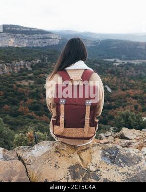 Rückansicht von jungen weiblichen Wanderer mit stilvollen Rucksack sitzend Auf felsigen Klippen und genießen Sie malerische Landschaft mit bunten Wald Und Berge im bewölkten Herbsttag Stockfoto