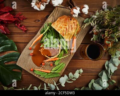 Overhead Wellington Sirloin serviert mit frischem Gemüse und Rotwein Auf Holztischplatte inmitten von Pflanzenzweigen Stockfoto