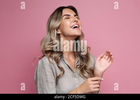 Studioaufnahme einer glücklichen emotionalen Frau mit lockigen Haaren, die weit lächelt, perfekte weiße Zähne zeigt, die Augen mit Vergnügen schließt, in Leinenhemd gekleidet, Stockfoto