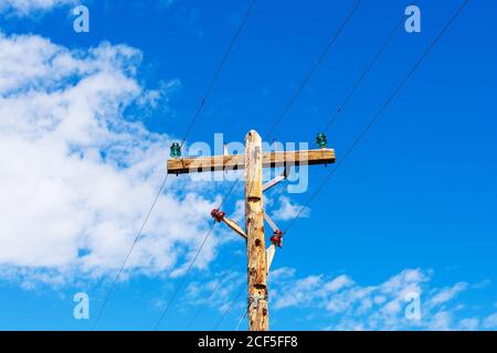 Verwitterter hölzerner Pfosten mit parallelen Einschaltungen und bunten Isolatoren am blauen Himmel. Stockfoto