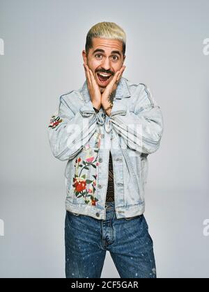 Junge ethnische Mann Grimasse überrascht Gesicht Blick auf die Kamera Tragen Sie eine trendige Denim-Jacke mit Blumenmuster, während Sie dagegen stehen Grauer Hintergrund Stockfoto