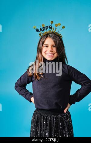 Happy preteen in schwarz und Gold Kopfbedeckung mit Wünschen glücklich Neujahr lächelt mit den Händen auf den Hüften gegen die Kamera Hellblauer Hintergrund im modernen Studio Stockfoto