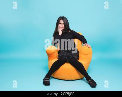 Präteen Mädchen in schwarzen Freizeitkleidung ruhende Hand auf Kinn Blick auf die Kamera, während Sie sich in hellgelben Sessel Hand entspannen Form vor hellblauem Hintergrund in zeitgenössischem Studio Stockfoto