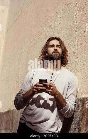 Junger bärtiger, hübscher Mann in beigem Pullover, der an der Wand gelehnt ist Und das Handy in den Händen halten, nachdenklich mitschauen Stockfoto