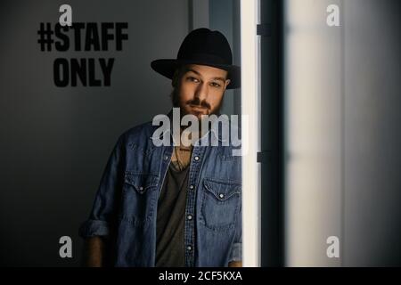 Junger bärtiger hübscher Mann in Hut und Jeanshemd suchen Bei der Kamera im dunklen Raum Stockfoto