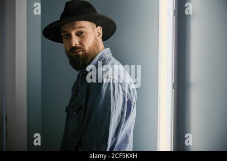 Junger bärtiger hübscher Mann in Hut und Jeanshemd suchen Bei der Kamera im dunklen Raum Stockfoto