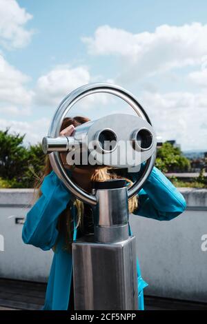 Anonyme blonde Frau in blauem Mantel, die durch Straßenmünze schaut Betrieben binokular, während auf Holzpflaster in der Nähe von Betonzaun stehen An sonnigen Tag Stockfoto