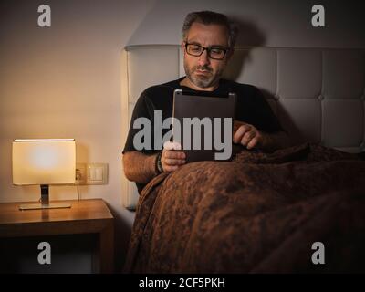 Entspannender Mann im Bett, der im Internet liest und ein Tablet vor dem Schlafen und Ausruhen Stockfoto