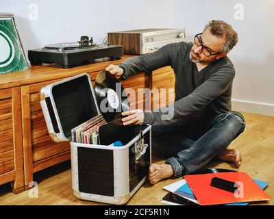 Stilvoller Mann in legerer Kleidung und Brille auf Holz sitzend Boden und Kommissionierung Vinyl-Platte, während das Wochenende zu Hause genießen Stockfoto