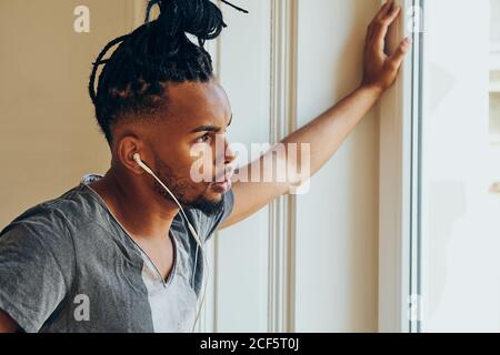 Seitenansicht eines ernsthaften ethnischen Mannes mit kreativer Frisur zuhören Zu Musik und lehnte sich auf Fensterrahmen Stockfoto