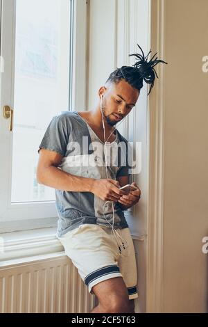 Junger afroamerikanischer Mann mit kreativer Frisur auf Fensterbank gelehnt Zu Hause mit dem Mobiltelefon und Musik hören In Ohrhörern Stockfoto