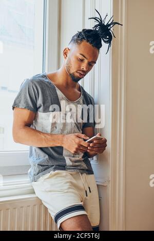 Junger afroamerikanischer Mann mit kreativer Frisur, der sich anlehnt Fensterbank und Surfen im Handy Stockfoto