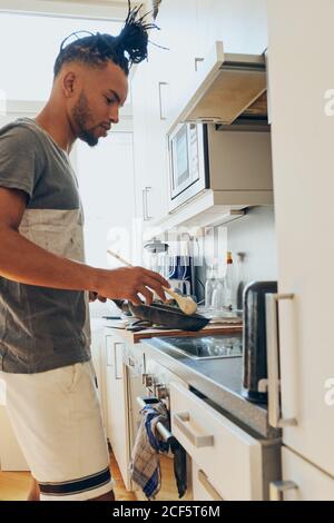 Seitenansicht des afroamerikanischen geflochtenen Mannes, der in der Küche stand Mit Bratpfanne und Scapula an sonnigen Tagen Stockfoto