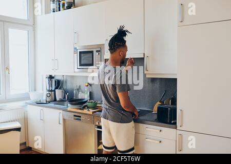 Seitenansicht des afroamerikanischen geflochtenen Mannes, der in der Küche stand Mit Bratpfanne und Scapula an sonnigen Tagen Stockfoto