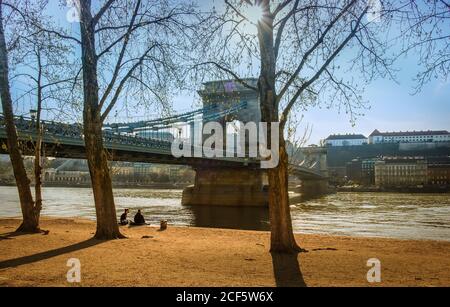 Budapest, Ungarn, März 2020, Blick auf ein junges Paar, das am Donauufer neben der Kettenbrücke von Szechenyi sitzt Stockfoto