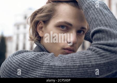 Moderne seriöse junge Männchen mit stilvollen Haarschnitt in grau warm Pullover berührt die Haare und blickt auf die Kamera, während er auf dem Pullover steht City Street Stockfoto