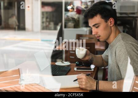 Seitenansicht des jungen Mannes mit Tasse Kaffee schreiben Im Notizblock, während man am Tisch hinter Glas sitzt und studiert In der Cafeteria Stockfoto