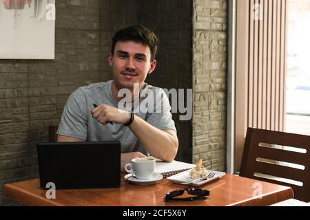 Junger Mann mit Laptop und Notebook lächelnd und anblickend Kamera beim Sitzen am Tisch mit Kaffee und Salat Gemütliches Restaurant Stockfoto