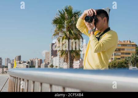 Inspirierter junger männlicher Fotograf im gelben lässigen Sweatshirt sprechend Foto Auf der Kamera, die im Sonnenlicht am Stadtkai steht Stockfoto