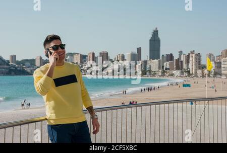 Zufriedener junger Mann in lässiger Kleidung und trendigen Sonnenbrillen reden Auf dem Handy am Wasser mit moderner Stadtlandschaft und Strand Im Hintergrund Stockfoto