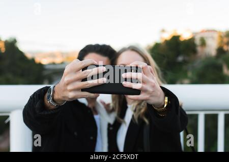 Fröhliche junge multirassische Paar in Freizeitkleidung unter Selfie an Mobiltelefon, während Sie zusammen auf einer Brücke mit grünen Bäumen stehen Und Stadtgebäude im Hintergrund Stockfoto