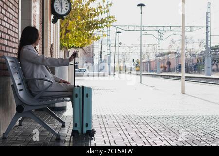 Seitenansicht der jungen stilvollen weiblichen Reisenden mit Koffer sitzen Auf der Bank und warten auf den Transport am Bahnhof in Sonniger Tag Stockfoto