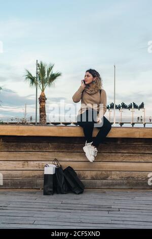 Zufriedene, lockige junge Frau in legerer Kleidung, die lächelt, während sie das Mobiltelefon auf der Bank mit Einkaufstaschen am modernen Kai benutzt Stockfoto
