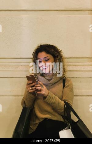 Zufriedene, lockige junge Frau in legerer Kleidung, die lächelt, während sie das Mobiltelefon mit Einkaufstaschen an der Wand anlehnt Stockfoto