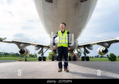Asiatischer Mann Ingenieur Wartung Flugzeug hält weißen Helm vor Flugzeug von Reparaturen, Fixes, Modernisierung und Renovierung im Flughafen. Stockfoto