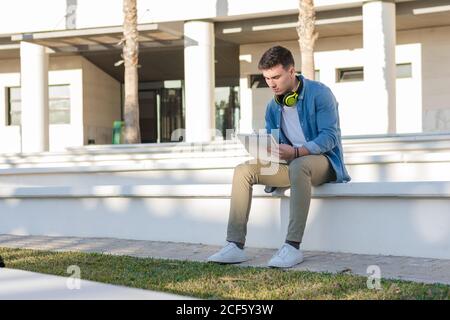 Nachdenklicher intelligenter männlicher Student in hellen Kopfhörern, der in der Universität studiert Quadrat und Schreiben in Notizbuch sitzt auf dem Zaun Stockfoto