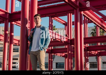 Smart stylish Mann mit Rucksack Blick auf Kamera stehen in Hof mit roten Metall-Konstruktion in sonnigen Tag Stockfoto