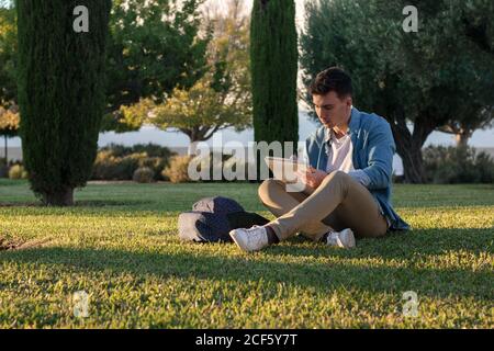 Smart konzentrierter Mann mit Rucksack studieren Schreiben auf Notizblock während Sitzen im Park Gras mit gekreuzten Beinen in sonnigen Tag Stockfoto