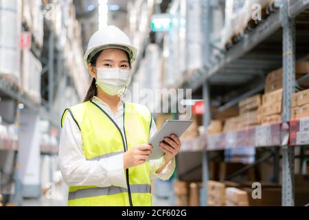 Junge asiatische Frau Auditor oder Trainee-Mitarbeiter trägt Maske während der COVID Pandemie im Lager Versand Industrie arbeiten. Looking up und Kontrollen Stockfoto