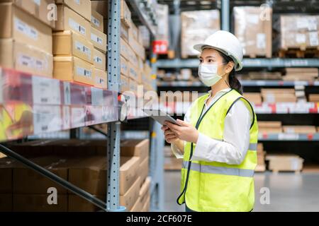 Junge asiatische Frau Auditor oder Trainee-Mitarbeiter trägt Maske während der COVID Pandemie im Lager Versand Industrie arbeiten. Looking up und Kontrollen Stockfoto